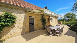 A stone building with a tiled roof, a covered patio with a table and chairs, and a barbecue grill.