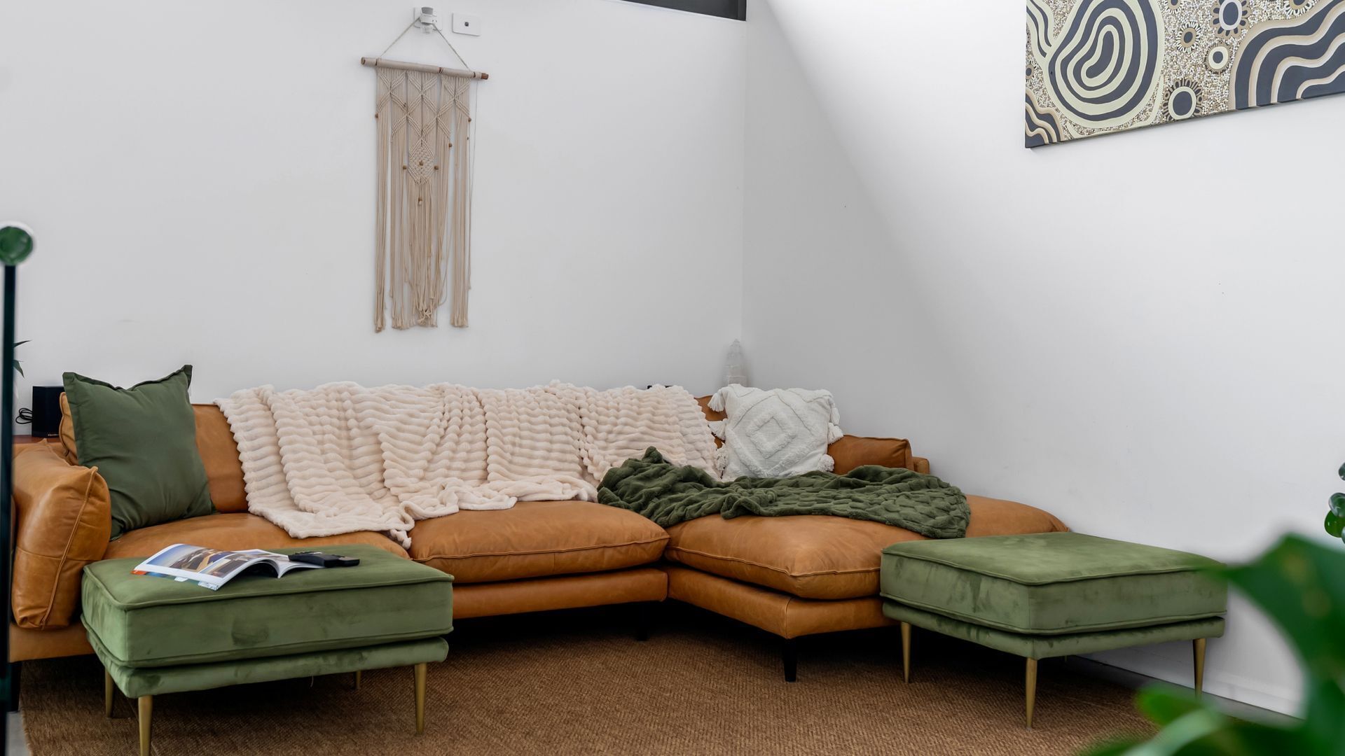 A cozy living room with a brown leather sofa, green and white throw blankets, and a macrame wall hanging.
