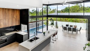 A modern kitchen with a large island, a dining area with outdoor seating, and a view of a swimming pool. - Belle Escapes Bluewater Breeze (Trinity Beach)