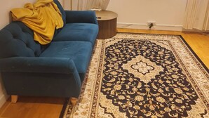 A living room with a blue sofa, a patterned rug, a lamp, and a view of buildings through a window. - 2 Beds Flat Centrally Located 2 Mins From Station (Romford)