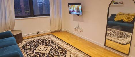 Una habitación con un ventanal grande, un sofá azul, un tapete estampado, un televisor montado en la pared y un espejo que refleja un sofá y un tapete.