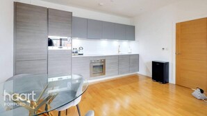 A modern kitchen with grey cabinets, a glass dining table, and a black speaker. - 2 Beds Flat Centrally Located 2 Mins From Station (Romford)