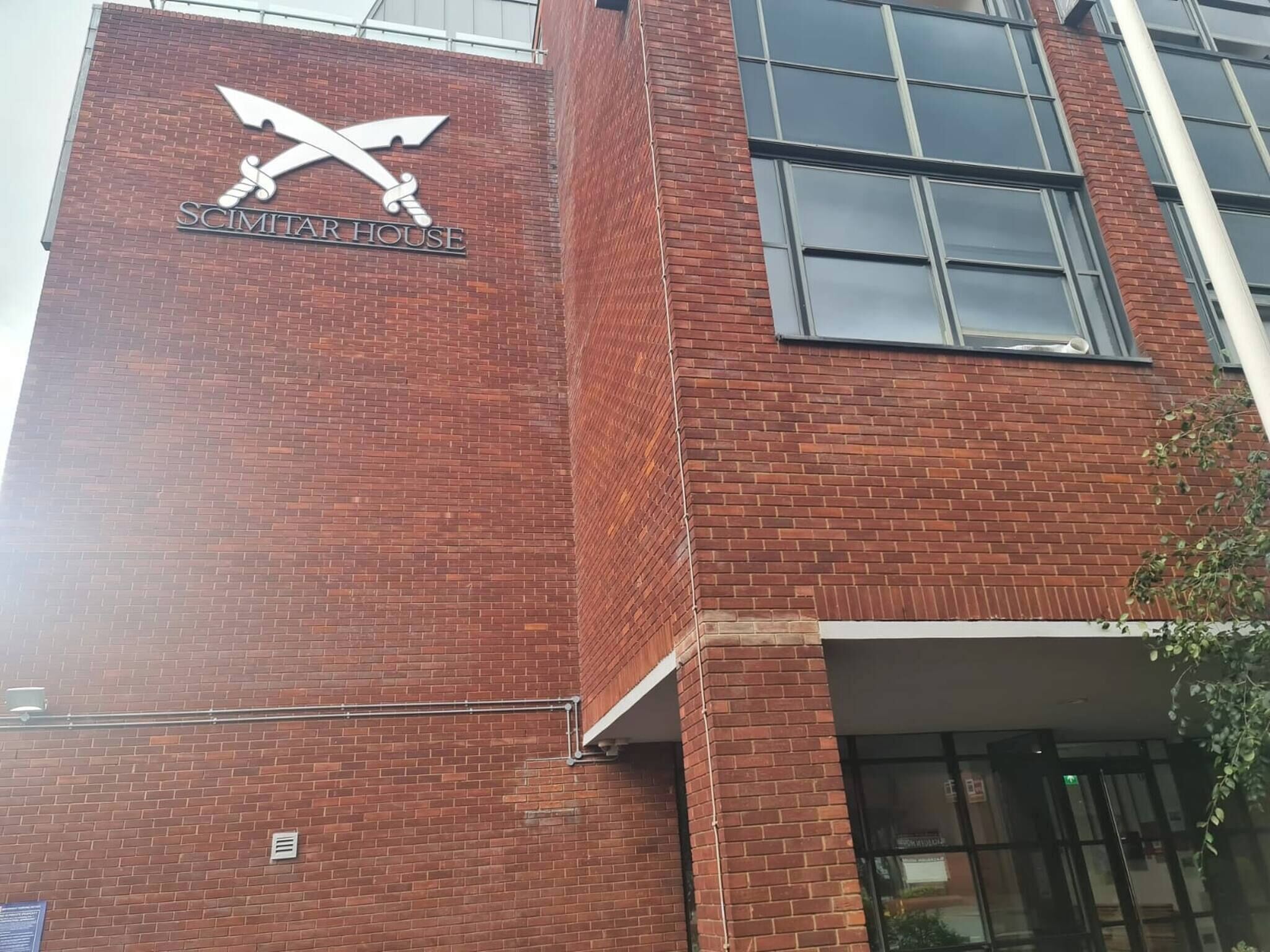 A brick building with a sign reading "SCIMITAR HOUSE" and a logo featuring crossed swords.