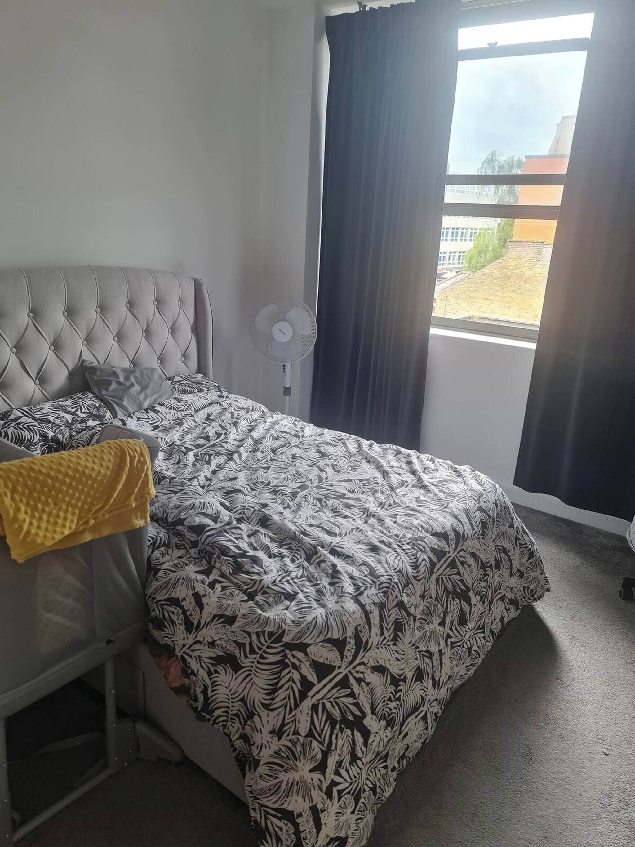 Un dormitorio con cabecera capitonada, cama con colcha estampada, manta amarilla, ventilador de pie y vistas a edificios a través de una ventana con cortinas.