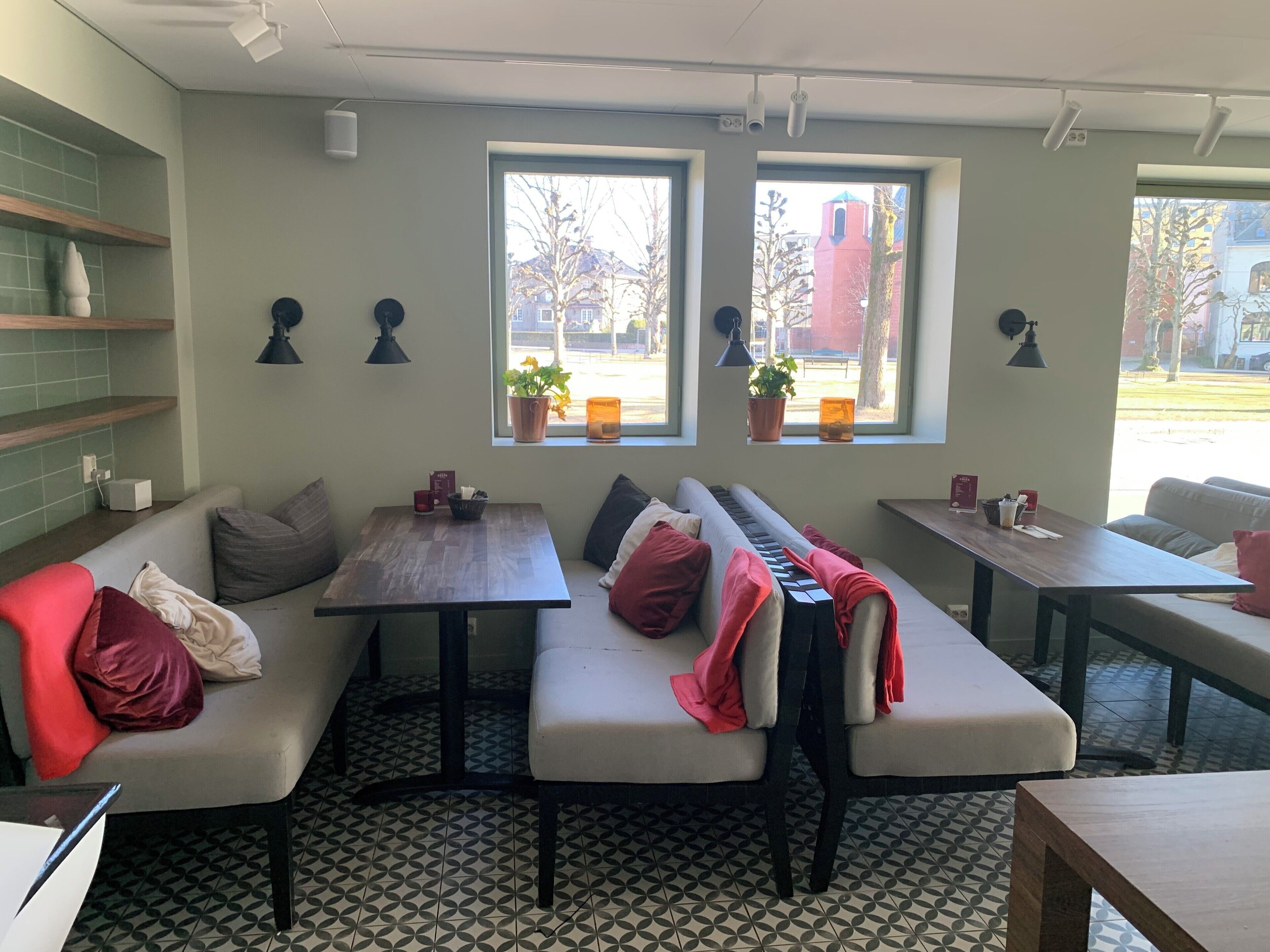 A cozy cafe with a long bench seating area, small tables, and potted plants by the windows.