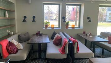A cozy cafe with a long bench seating area, small tables, and potted plants by the windows.