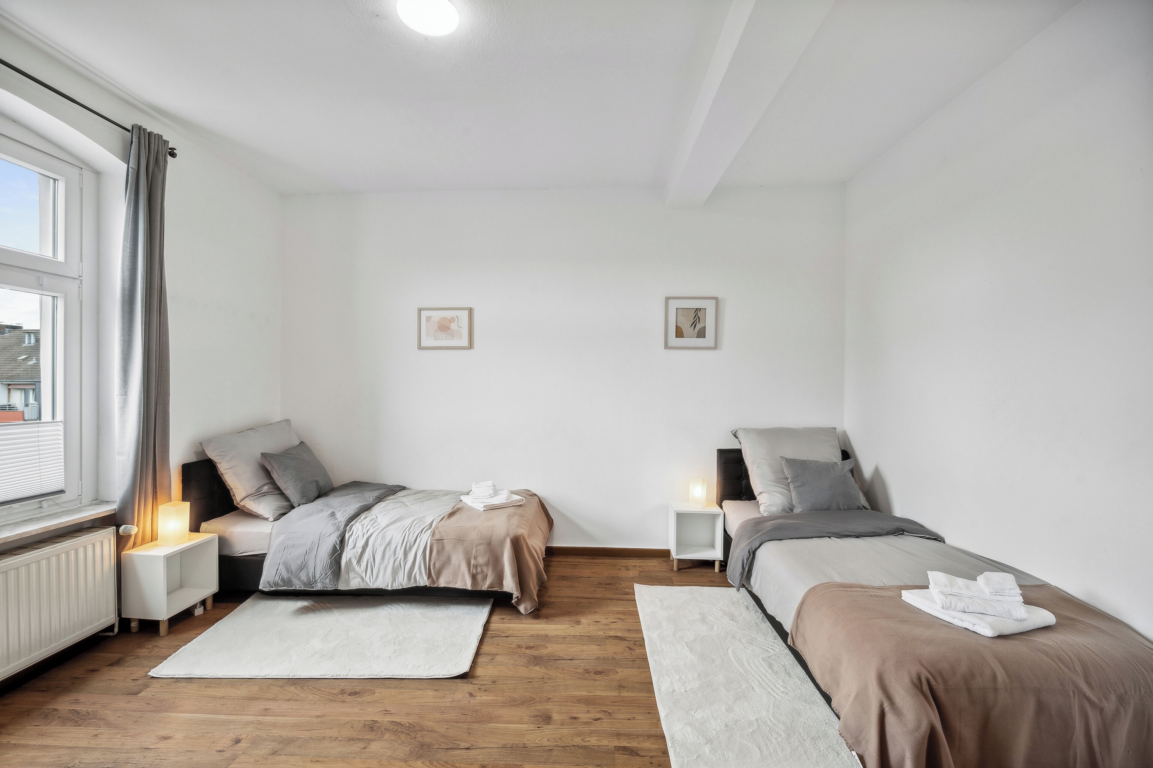 Ein Schlafzimmer mit zwei Betten, einem Fenster mit Vorhängen, Nachttischlampen und Bildern an den Wänden.