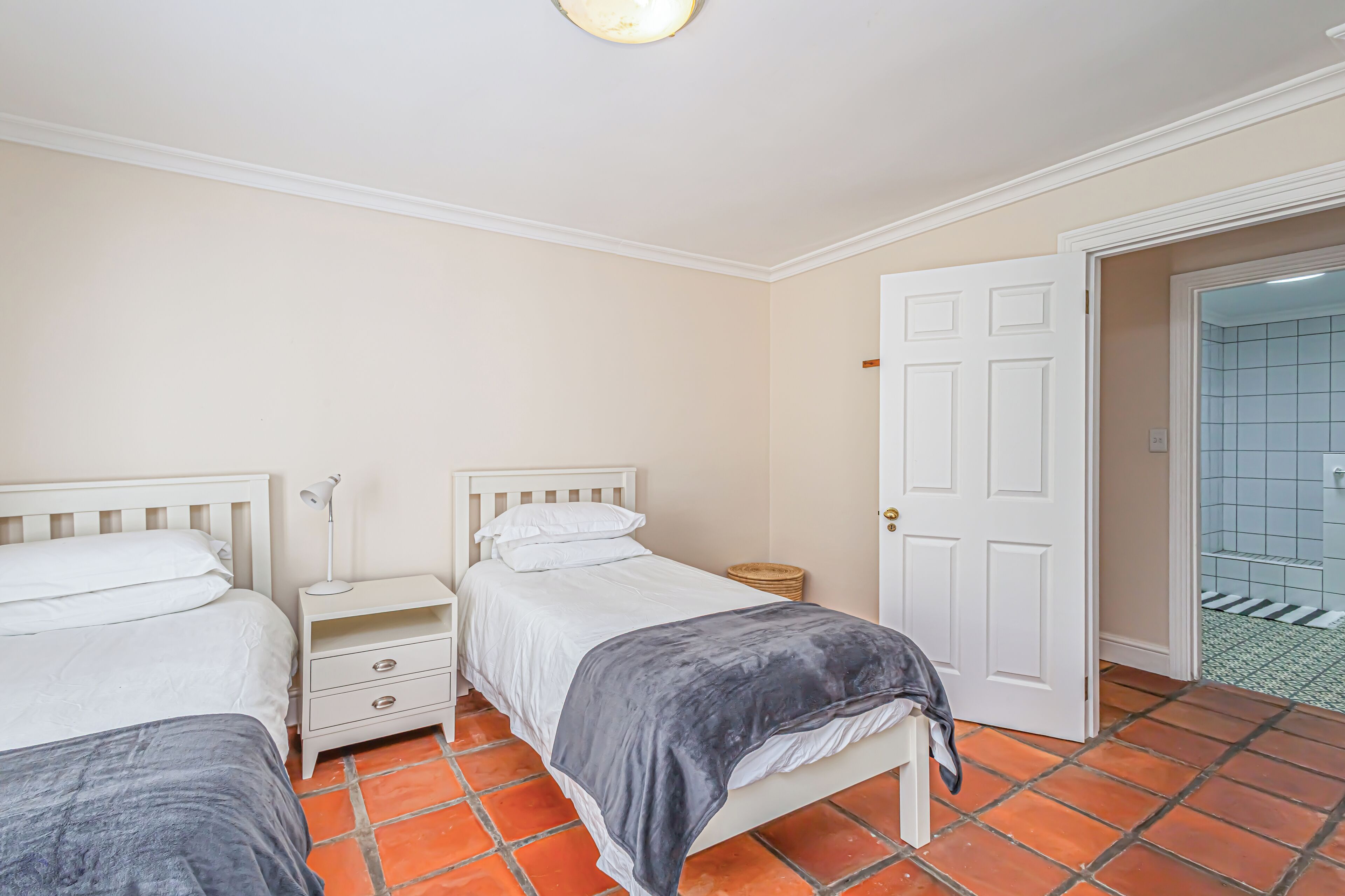 Een slaapkamer met twee bedden, een nachtkastje, een deur naar een badkamer en een betegelde vloer.