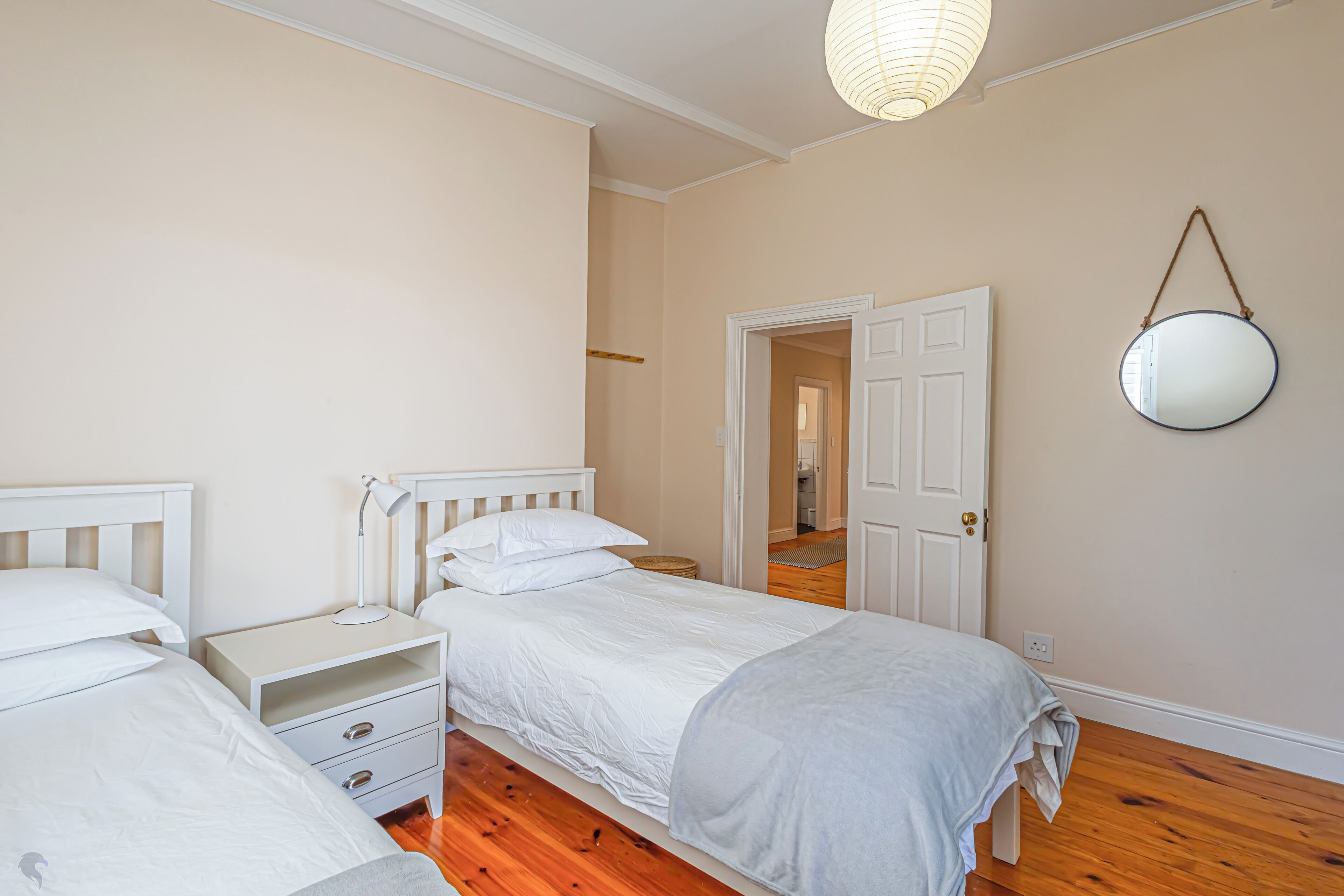 A bedroom with two single beds, a bedside table with a lamp, a mirror on the wall, and a door leading to another room.