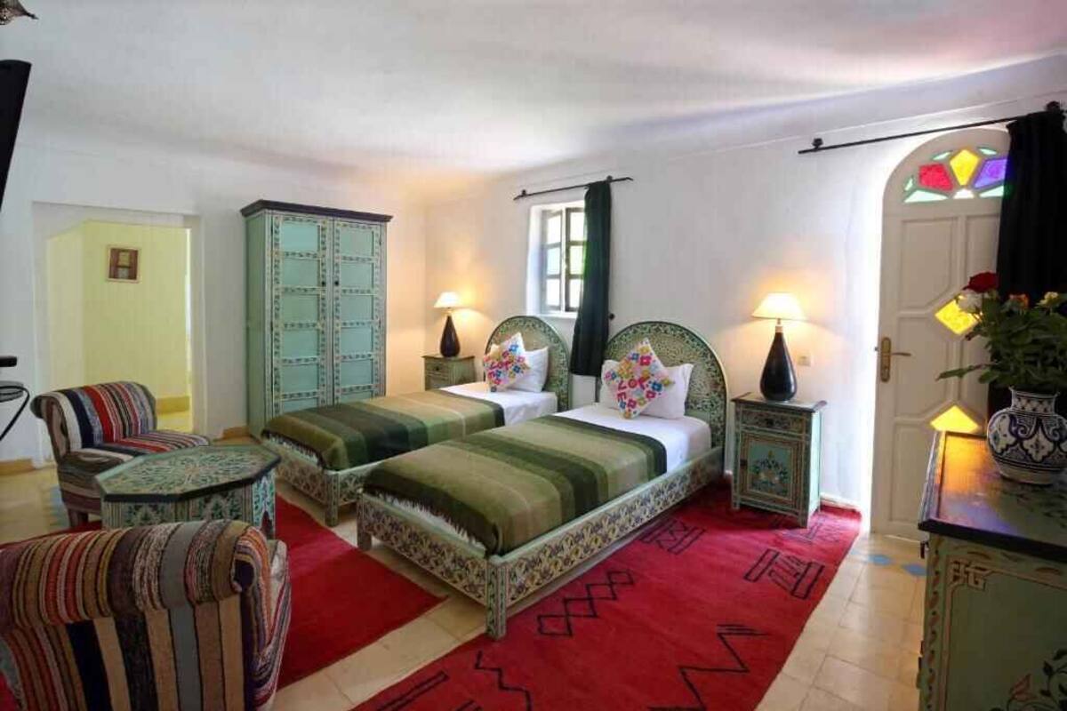 Une pièce avec deux lits, une moquette rouge, des chaises à rayures, un buffet et une petite table.
