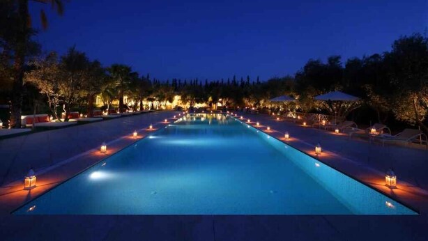 A nighttime view of a swimming pool with surrounding lounge areas and trees. - Le Bled (Marrakech)