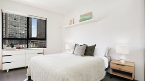 A bedroom with a large bed, a nightstand, a dresser, and a window with a city view. - RARE FIND! Modern 2 Bdrm, 2 Bath, 1 Car in Central Sydney (City of Sydney)