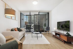 A modern living room with a sofa, coffee table, and TV. There is a sliding glass door leading to a balcony with outdoor furniture. - RARE FIND! Modern 2 Bdrm, 2 Bath, 1 Car in Central Sydney (City of Sydney)
