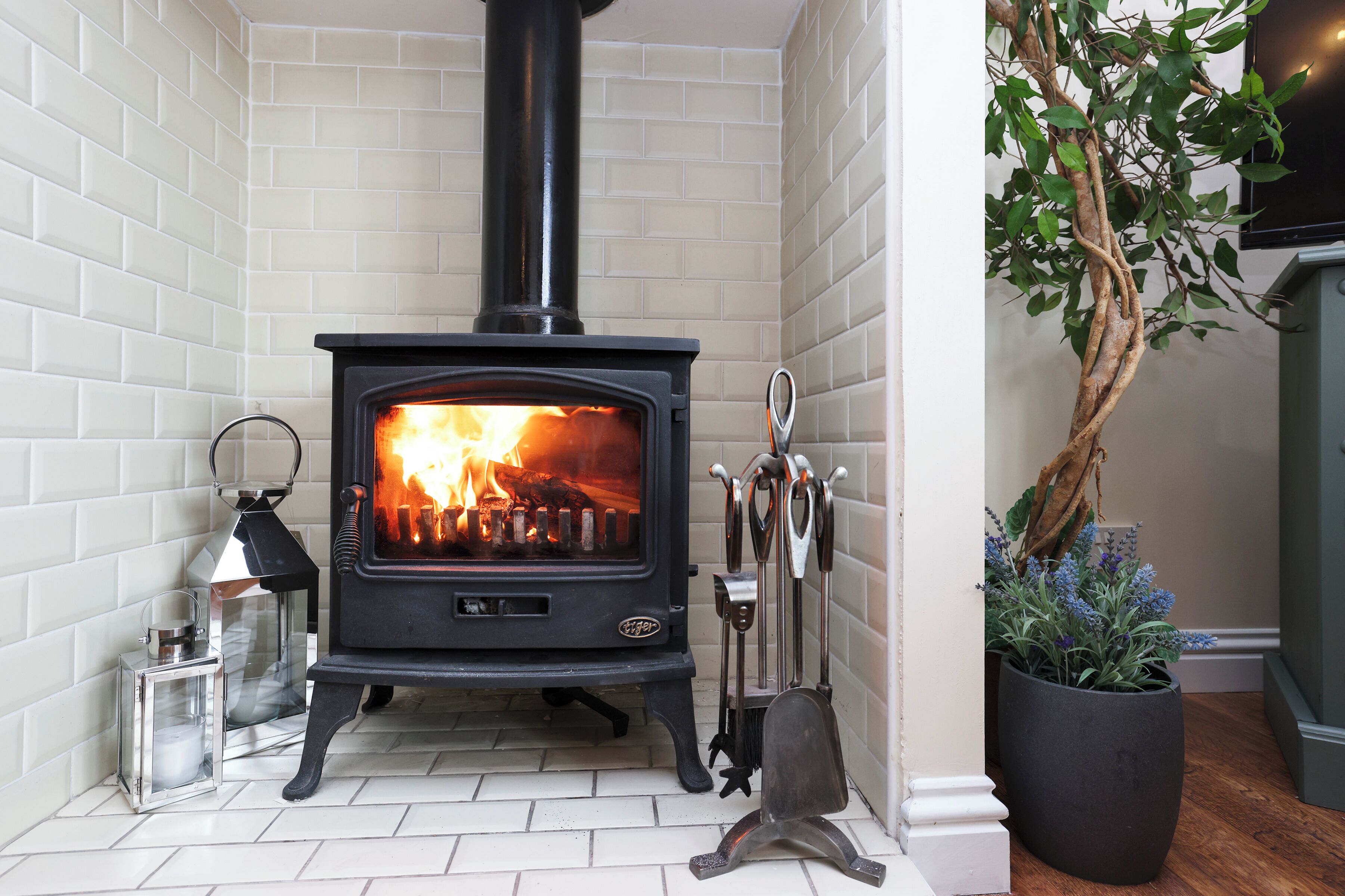 A wood-burning stove in a corner with a white tiled wall, a potted plant, and a lantern.