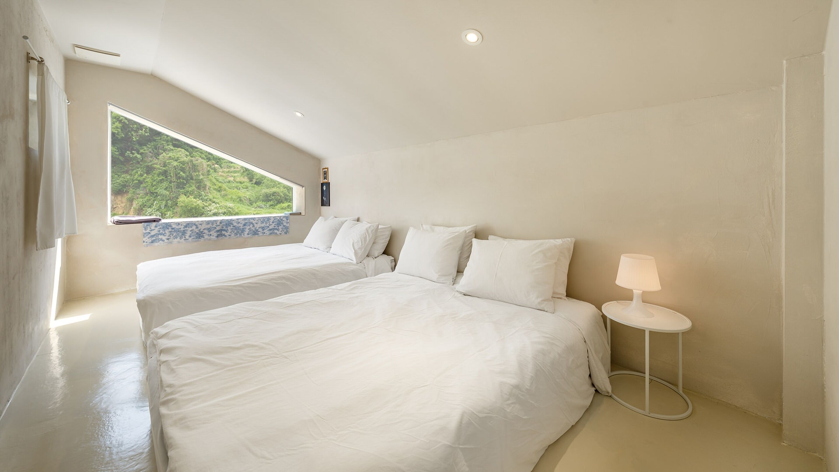 A bedroom with two beds, a window with a view of greenery, a small bedside table with a lamp, and a ceiling light.
