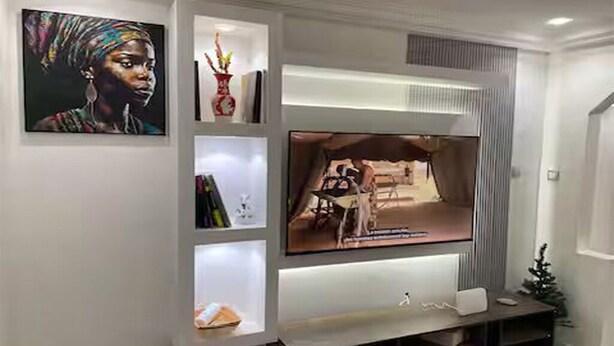 Uma sala de estar moderna com uma TV de tela plana fixada na parede, cercada por estantes com diversos objetos decorativos.