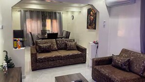 Uma sala de estar com um sofá marrom, uma mesa de centro e uma televisão. Há um quadro na parede e um aparelho de ar condicionado.