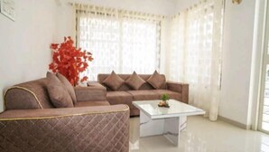 A modern living room with a brown sectional sofa, a glass coffee table, and a large window with sheer curtains. - Saara Residency (Pune)