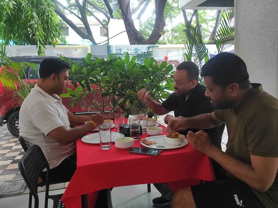 A group of people dining at an outdoor table with a red tablecloth, surrounded by greenery.