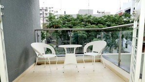 Een balkon met twee witte wickerstoelen en een klein rond tafeltje, met een glazen reling en uitzicht op groen en gebouwen.