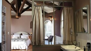 Un dormitorio con cama, una silla, un ventanal con contraventanas y un baño con lavabo y espejo.