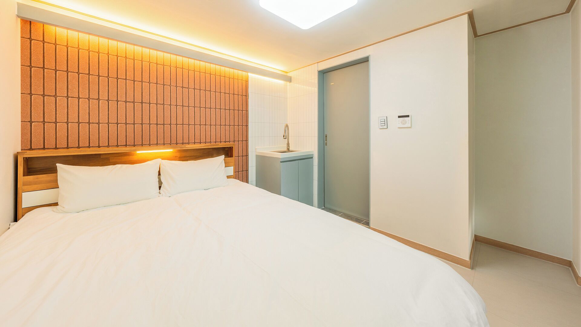 Habitación de hotel con una cama grande, cabecera de madera, estante empotrado, baño pequeño con lavamanos y una puerta.