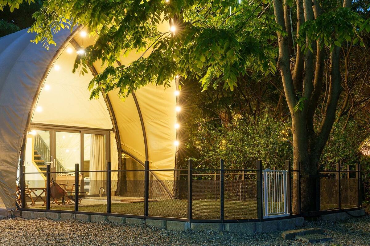 A large, semi-transparent dome tent with a covered patio area, surrounded by trees and a gravel ground.