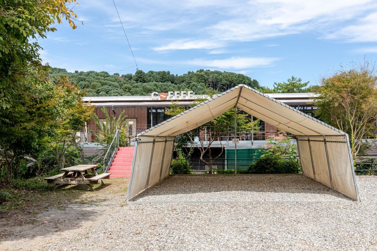 Een grindpad met een houten constructie die op een tent lijkt, een gebouw met een bord waarop 'Café' staat, en een rode trap die naar een deur leidt.