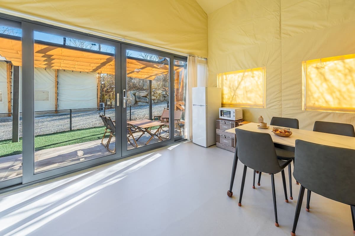 A modern, well-lit room with a dining table, chairs, and a kitchen area with a refrigerator and microwave. Large windows provide a view of the outdoors.