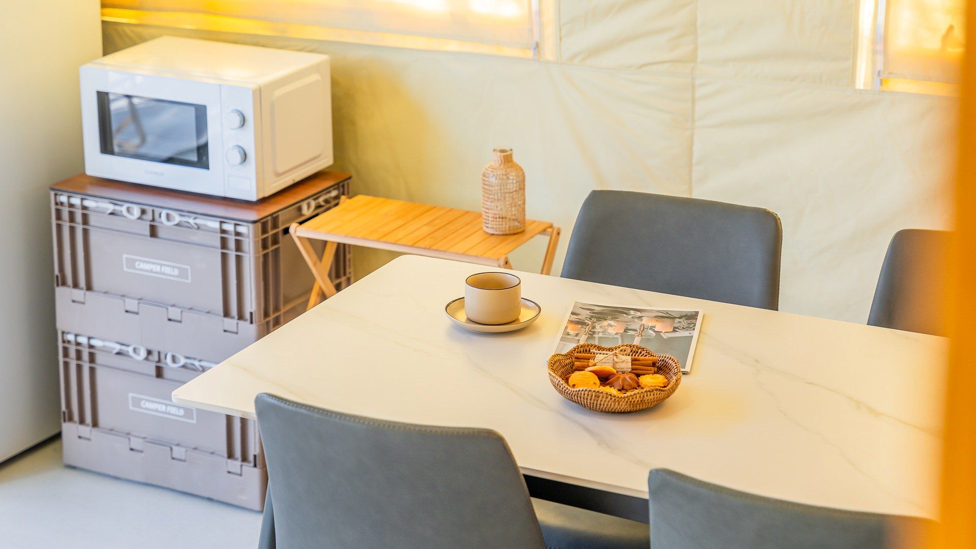 A compact dining area with a white table, grey chairs, a small wooden side table, a white microwave, and a basket of pastries.