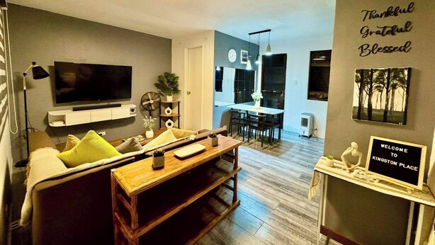 A modern living room with a TV, sofa, and coffee table. There's a kitchen in the background with a dining area. - Cozy Holiday Home in San Pablo City for Groups of 8-10 (San Pablo)