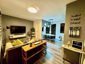 A modern living room with a TV, sofa, and coffee table. There's a kitchen in the background with a dining area.