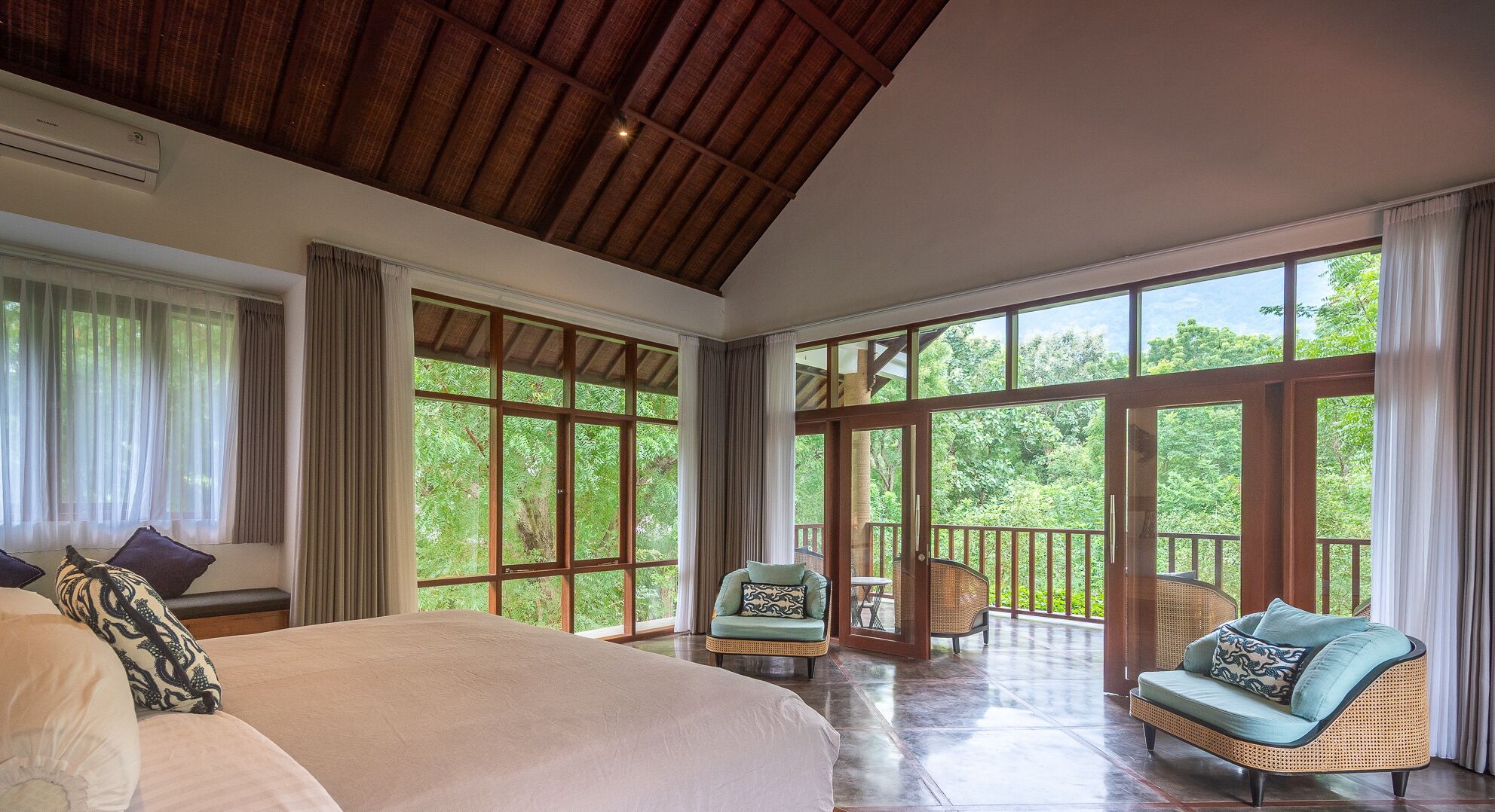 A spacious bedroom with a large bed, a balcony with wicker furniture, and large windows offering a view of the greenery outside.