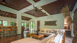A spacious living area with a high ceiling, wooden beams, a ceiling fan, and a large window providing natural light. - Luxury Living in Harmony with Nature! (bali)