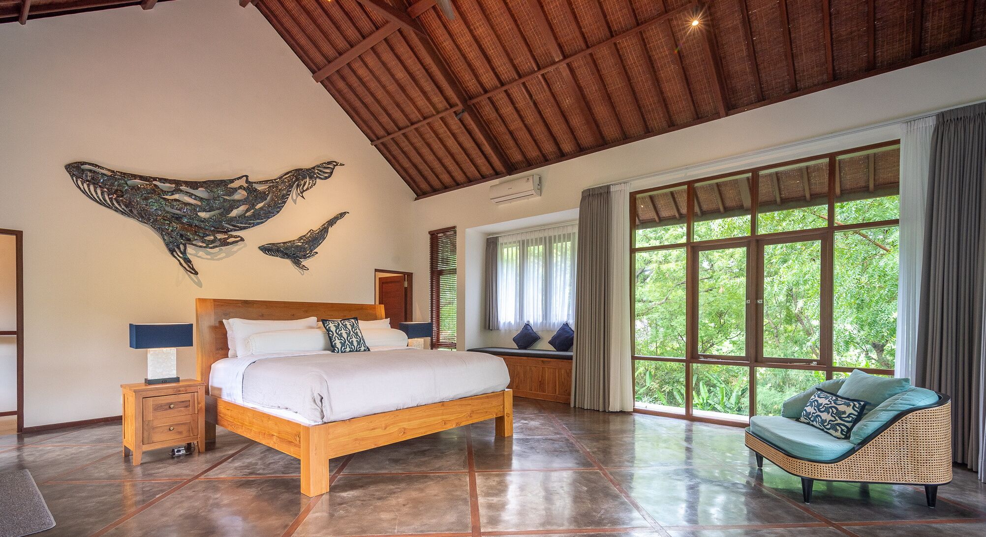A spacious bedroom with a large bed, a rattan chair, and a view of greenery through large windows.