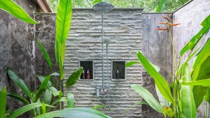 A stone wall with two small windows, a metal showerhead, and surrounding lush green foliage. - Luxury Living in Harmony with Nature! (bali)