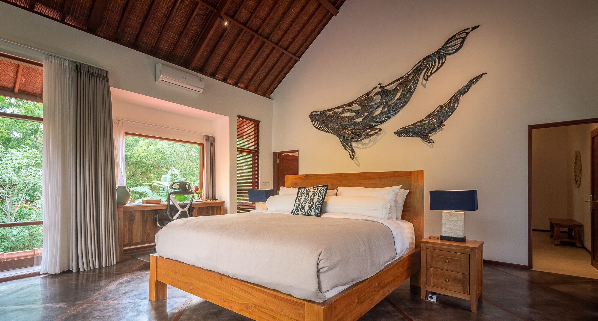 A spacious bedroom with a large bed, a desk, and a chair, a wooden ceiling, and a view of greenery outside.