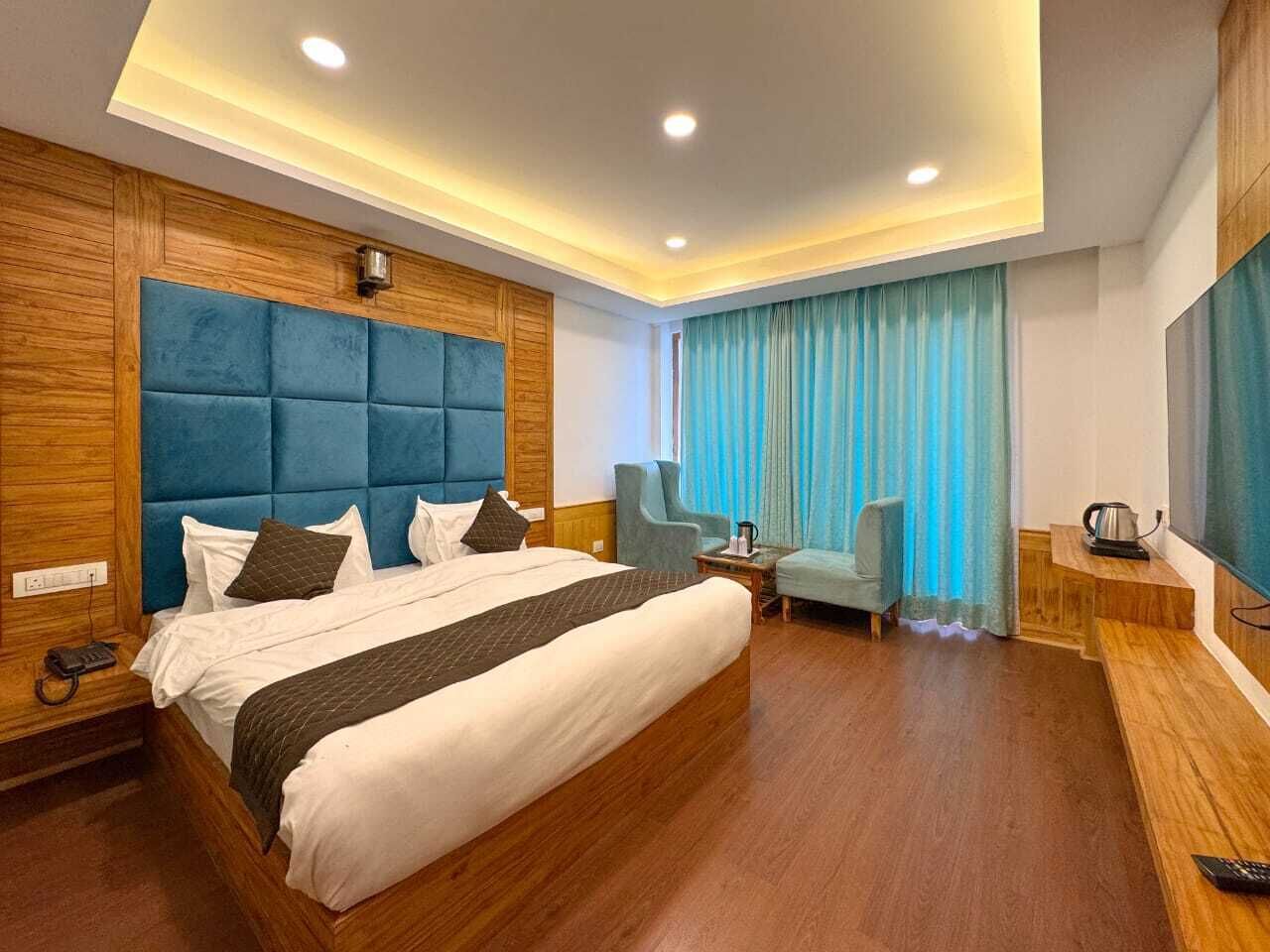 Habitación de hotel moderna con una cama grande, cabecera de madera, zona de estar con una silla, una pequeña mesa y un televisor.
