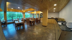 Un restaurante con mesas y sillas de madera, una zona de cocina con electrodomésticos de acero inoxidable y grandes ventanales con vistas a árboles.