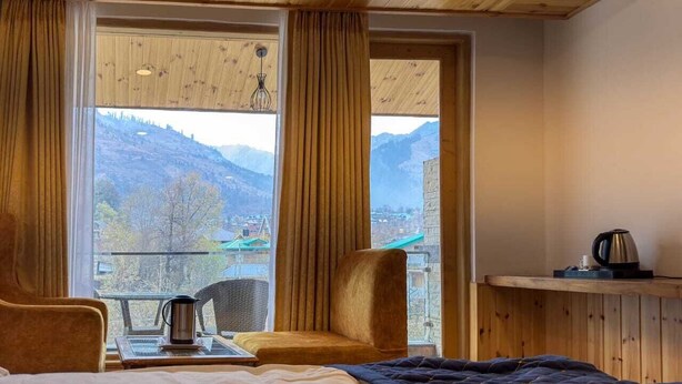 Una habitación acogedora con techo de madera, un ventanal con cortinas, una silla, una mesa con hervidor y vistas a las montañas.