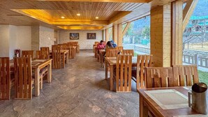 Comedor con paneles de madera, mesas y sillas, suelo de piedra y grandes ventanales que ofrecen vistas al exterior.