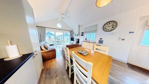 A cozy room with a wooden dining table, white chairs, a brown sofa, and a view of a blue house through a glass door. - Badger's Retreat -  a beach house that sleeps 4 guests  in 2 bedrooms (Filey)