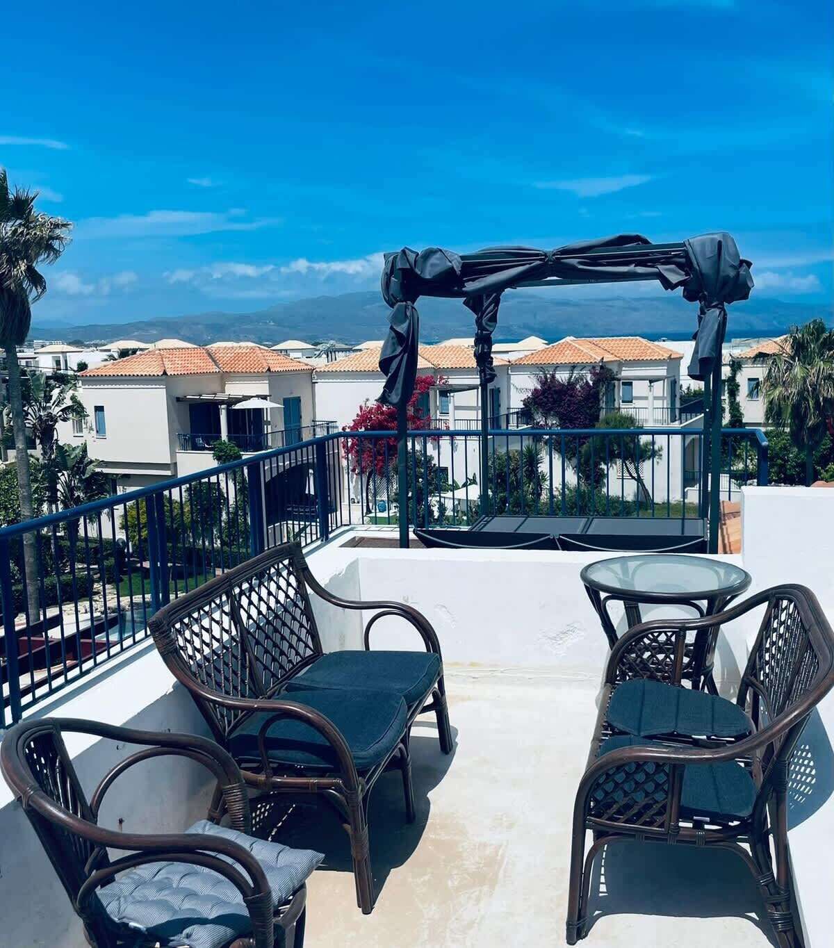 A terrace with wicker furniture and a pergola, overlooking a coastal town.
