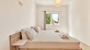 A minimalist bedroom with a wooden bed, a bedside table, a hanging light fixture, and a window with a view of greenery. - Cassiopeia Penthouse (Pyrgos Psilonerou)