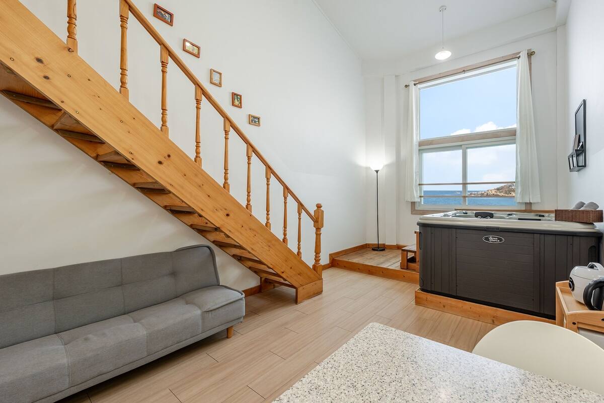 A modern living room with a wooden staircase, a gray sofa, a kitchen area with a hot tub, and a large window overlooking the sea.