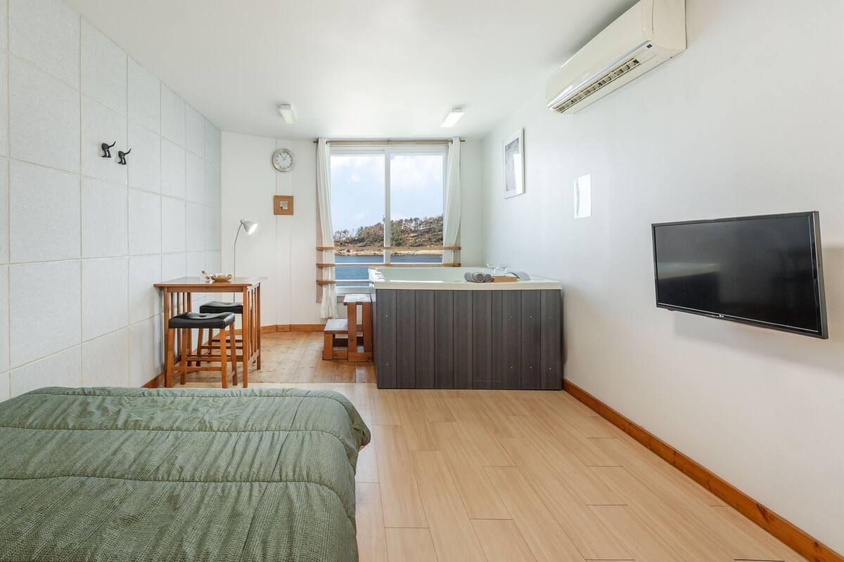 A compact room with a bed, a desk with a chair, a small kitchen area, and a TV mounted on the wall.