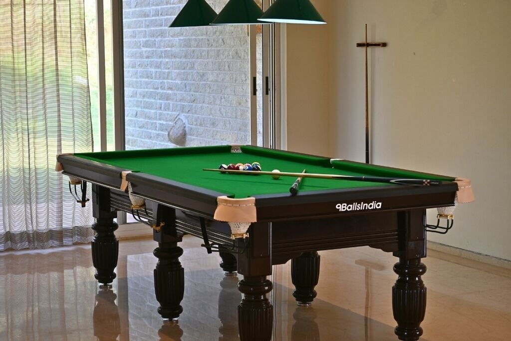 A pool table with a green felt surface, branded with "9BallsIndia," in a room with a brick wall and a window with sheer curtains.