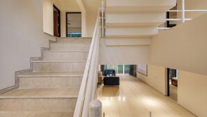 Un interior moderno con una escalera de mármol que conduce a un segundo piso, una zona de estar con sofá y una mesa de centro, y un ventanal que proporciona luz natural.