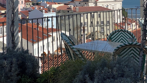Blick von einem Balkon mit einer Pergola auf eine Küstenstadt mit historischen Gebäuden und einem markanten Kirchturm.