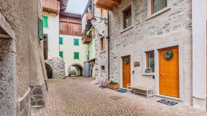 Una calle empedrada flanqueada por edificios, con muros de piedra, balcones de madera y un pequeño banco.